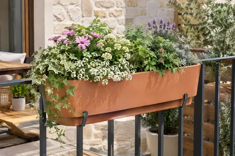 Balconnière terre cuite à support intégré, garnie de fleurs et installée sur une rambarde de balcon.